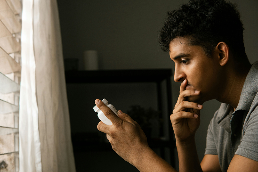 Man sitting by a window, staring at a pill bottle in his hand with concern, symbolizing the struggles that lead to opioid rehab treatment.
